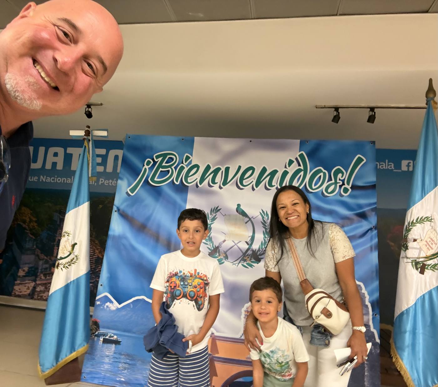 A family taking a selfie in front of the ¡Bienvenidos! Guatemala sign at La Aurora airport arrivals