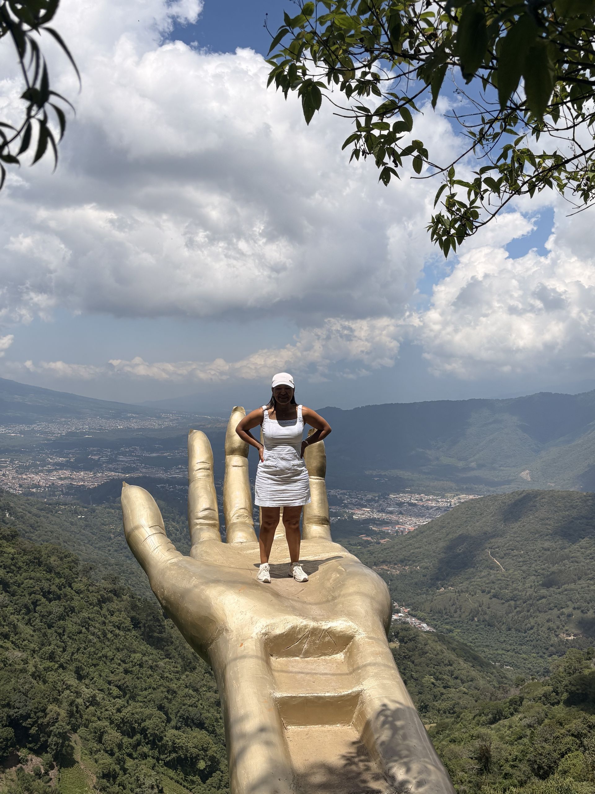 Visitor standing on the giant golden hand sculpture at Hobbitenango, high above the Antigua valley with volcano views
