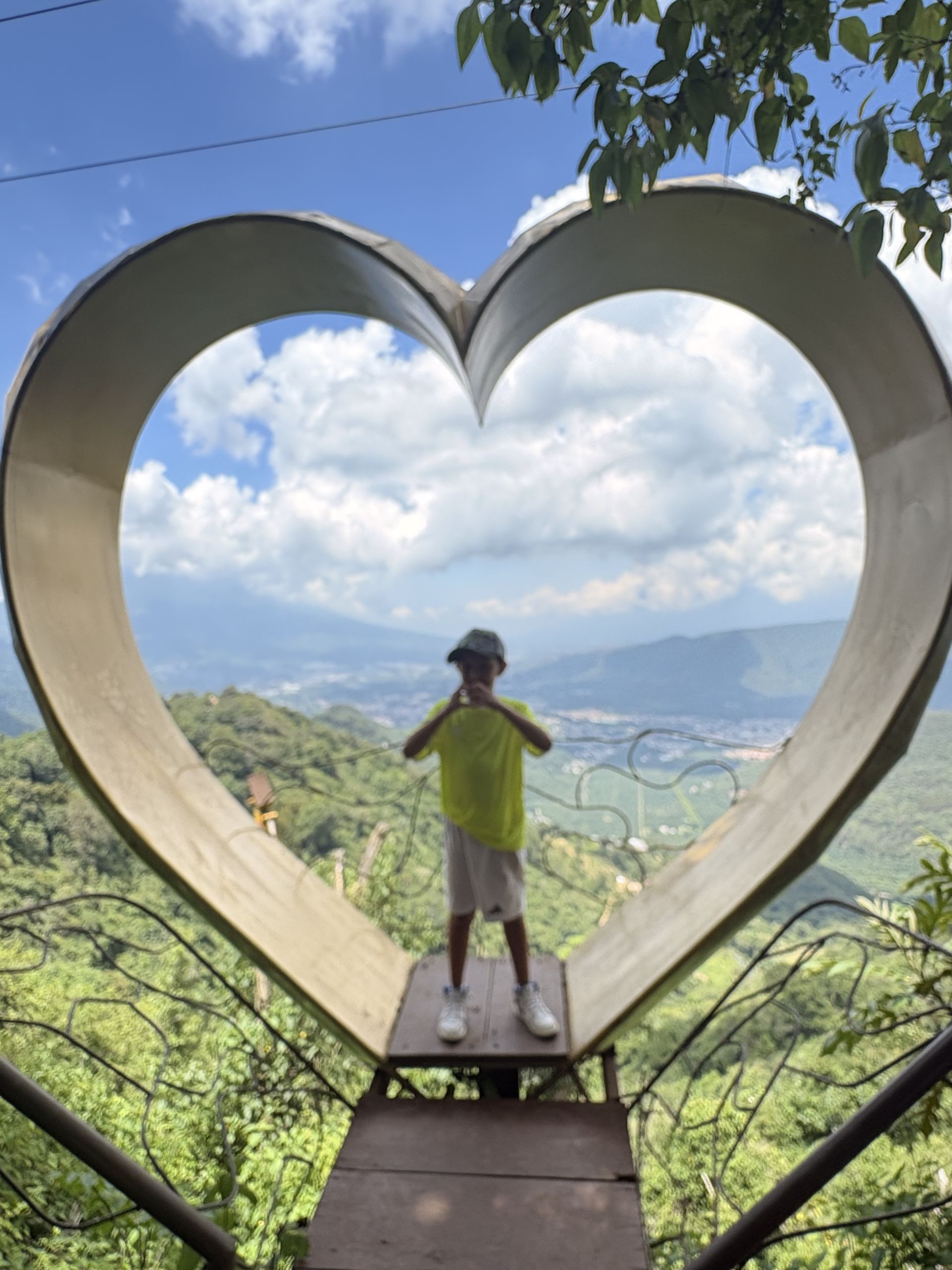 Child standing inside the giant heart-shaped frame at a Hobbitenango viewpoint overlooking the valley below