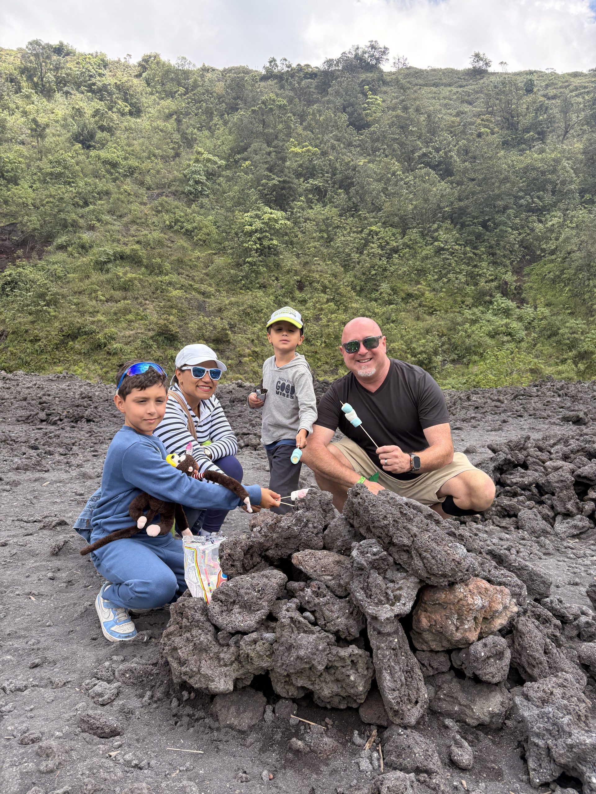 Family roasting marshmallows over hot volcanic rock on Pacaya volcano, green slopes rising behind them